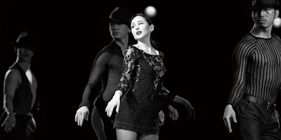 Four dancers perform on stage in black attire, with the central woman in a lace dress highlighted, all striking expressive poses under dramatic lighting.