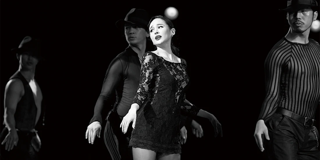 Four dancers perform on stage in black attire, with the central woman in a lace dress highlighted, all striking expressive poses under dramatic lighting.