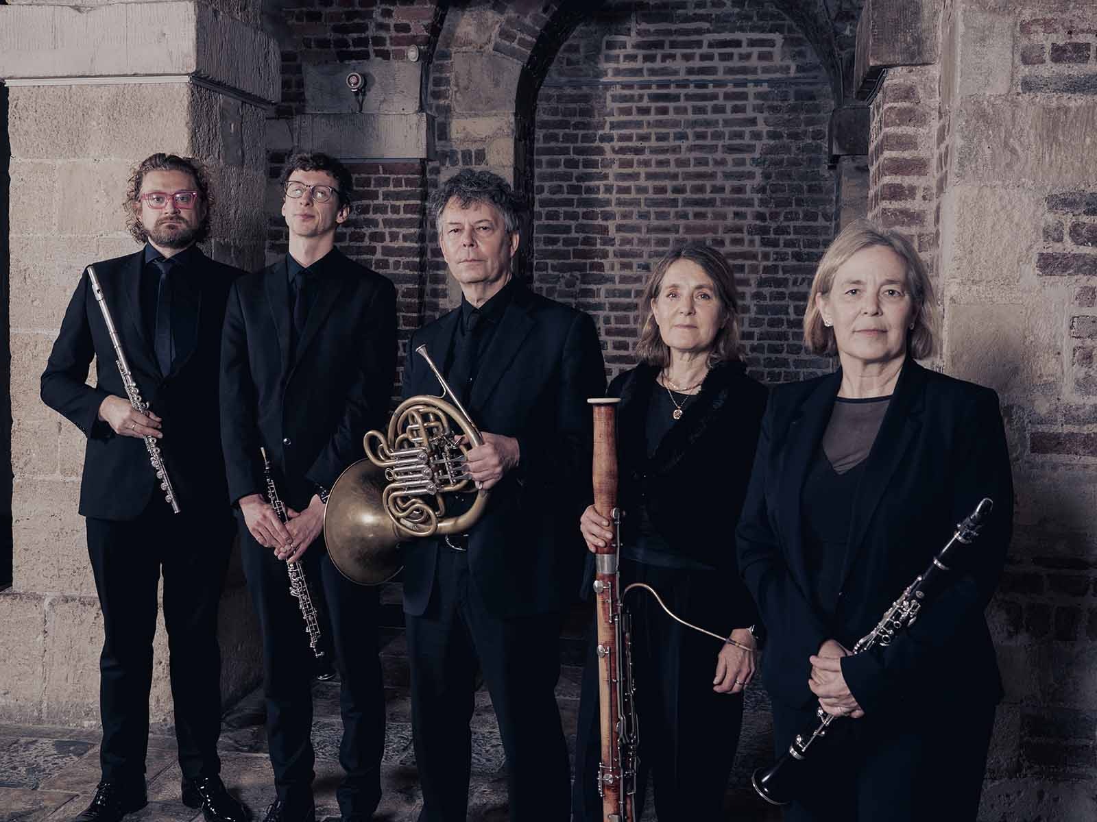 The ensemble in black formal attire stand indoors holding flute, French horn, bassoon, and clarinet, against a backdrop of stone and brick arches.