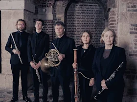 The ensemble in black formal attire stand indoors holding flute, French horn, bassoon, and clarinet, against a backdrop of stone and brick arches.