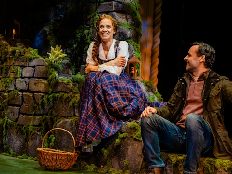 A woman in a plaid skirt sits on a mossy stone stage set, smiling, with a basket by her feet. A man sits beside her, looking at her.