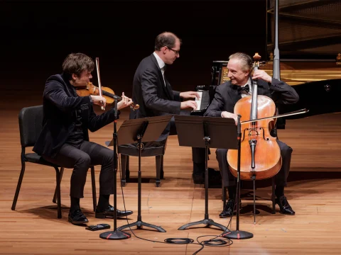 Three musicians perform onstage: a violinist, a pianist, and a cellist. They are positioned near each other, with music stands in front of the violinist and cellist.