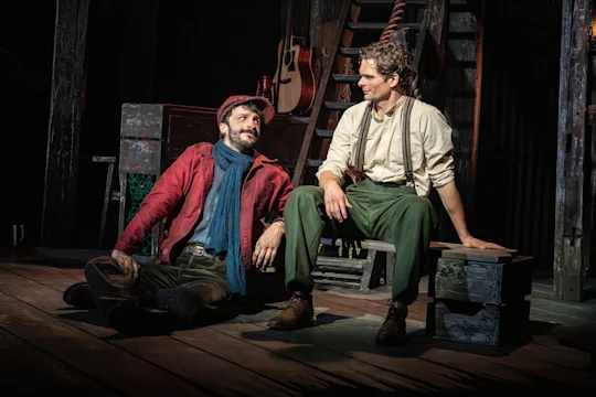 Two men sit on a dimly lit wooden stage, one leaning against a crate and the other perched on a bench, looking toward each other.