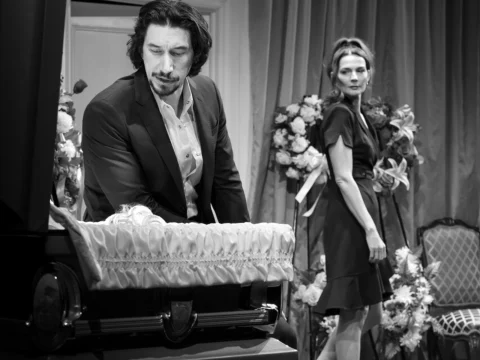 A man in a suit stands beside a coffin with flowers, while a woman in a dress looks on from behind in a black and white setting.