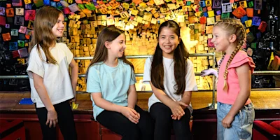 Four young girls smiling and talking together in front of a colorful wall decorated with alphabet blocks.