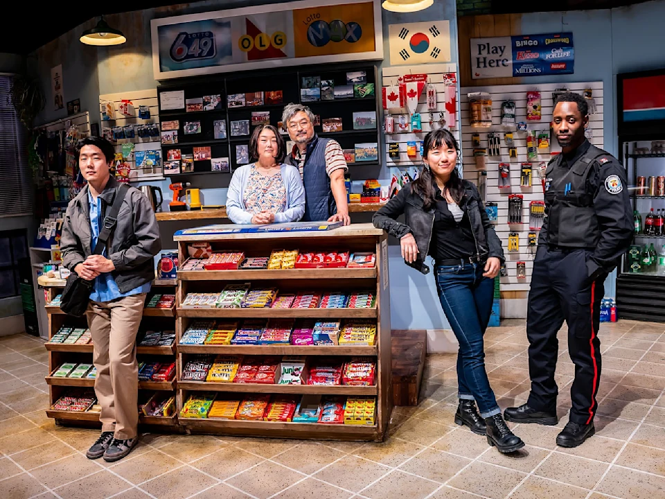 Five people pose inside a convenience store, standing around a counter and candy display, with lottery signs, flags, and various items visible in the background.