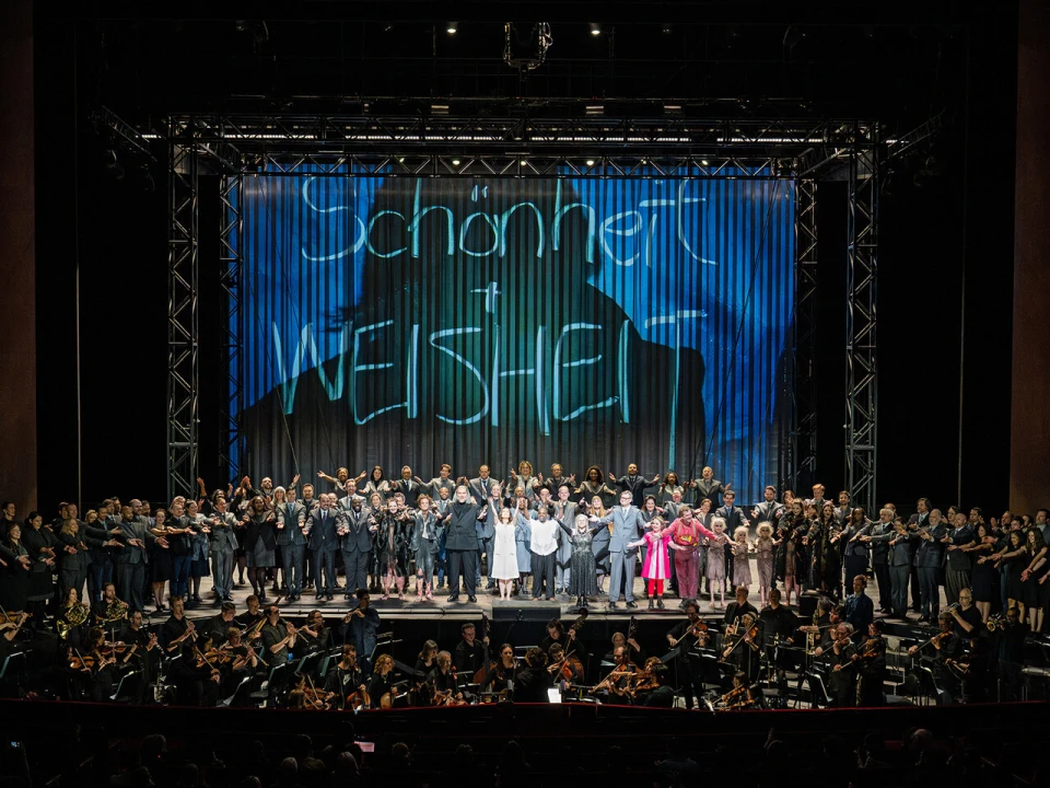 A large group of performers stand on stage with hands joined, facing the audience. They are on a set with a backdrop displaying the words "Schönheit und Wahrheit." An orchestra is seated in the pit below.