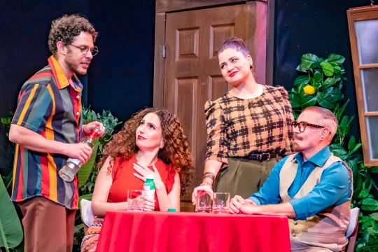 Four adults are gathered around a table with a red tablecloth. One man stands pouring a drink while the other three sit. They are indoors with a door and plants in the background.