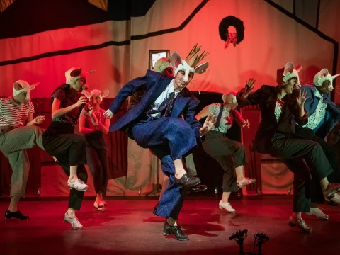 Performers dressed as mice in suits and casual attire dance energetically on a stage, bathed in red lighting, with a backdrop of a simple house outline.