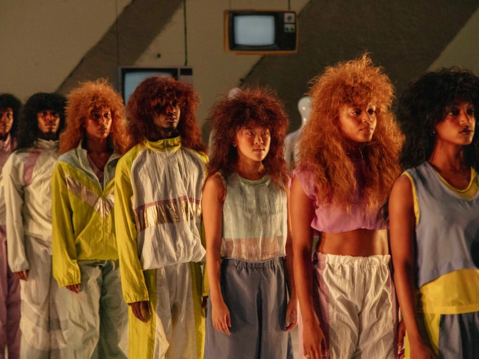 Production photo of A.I.M by Kyle Abraham: Cassette Vol. 1 in New York, showing A group of models with voluminous hairstyles stand in two lines, wearing pastel and neon-colored outfits, in an indoor setting with dim lighting.