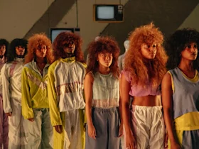 Production photo of A.I.M by Kyle Abraham: Cassette Vol. 1 in New York, showing A group of models with voluminous hairstyles stand in two lines, wearing pastel and neon-colored outfits, in an indoor setting with dim lighting.