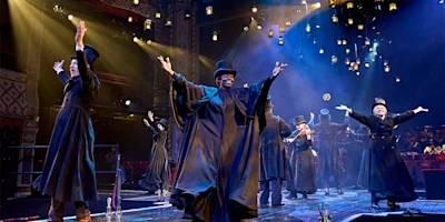 A group of actors in black robes and top hats perform energetically on a theater stage under dramatic lighting with an audience visible in the background.