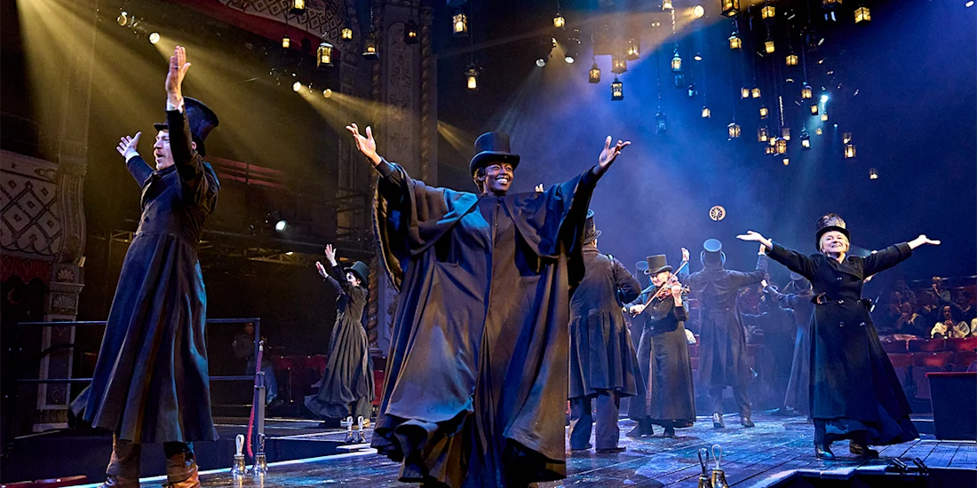 A group of actors in black robes and top hats perform energetically on a theater stage under dramatic lighting with an audience visible in the background.