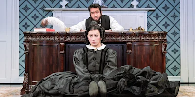 A woman in a black dress sits on the floor in front of an ornate desk, while a man sits behind the desk looking at her. The room has patterned wallpaper and formal decor.