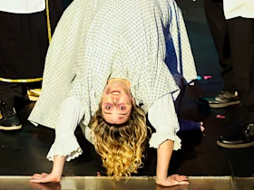 Person in a patterned dress performs a backbend on stage, facing the camera, with others standing nearby out of full view.