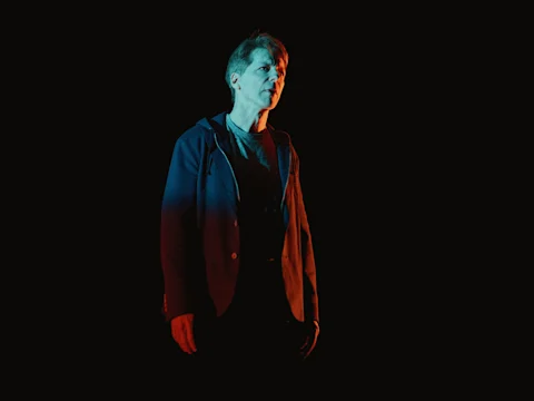 Production photo of The Unknown showing a person standing against a dark background, illuminated by dramatic red and blue lighting, wearing a jacket and looking forward.