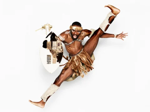 A person dressed in traditional attire performs an energetic high kick while holding a shield and spear.