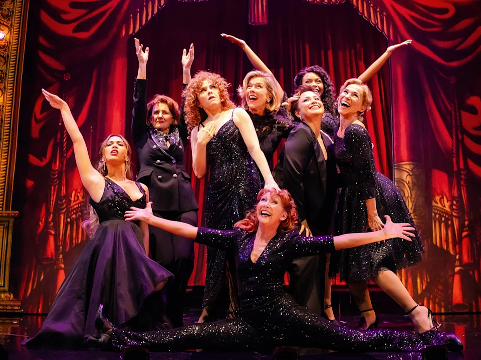 A group of nine individuals, predominantly women, strike a dynamic pose on stage in front of a vibrant red curtain, all dressed in elegant black attire.