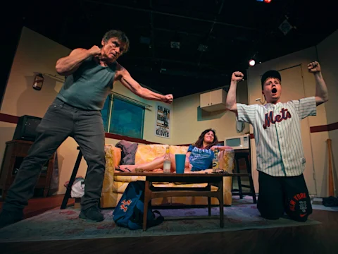 Three people are on a stage set designed as a living room; two men stand raising their arms while a woman sits on a couch between them, watching.
