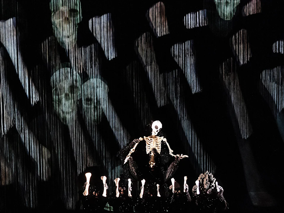 A person in a skeleton costume sits on stage with skeletal props, surrounded by projected skull and bone patterns in the background.