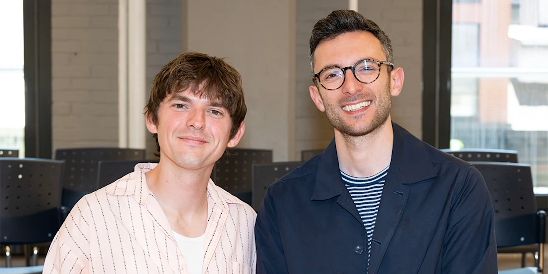 Two men are smiling and posing for a photo indoors; they are seated with empty chairs and large windows in the background.