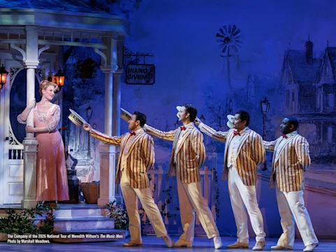 A woman stands on a porch while five men in striped blazers and boater hats sing to her onstage during a theatrical performance.