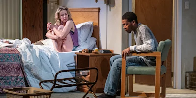A woman sits on a bed looking concerned while a man sits on a chair facing away from her in a cluttered motel room.