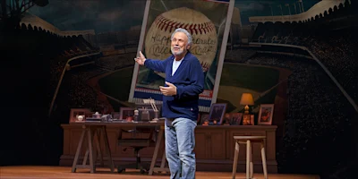 An older man in a cardigan stands on stage in front of a desk and a large oversized baseball card backdrop, gesturing toward the card.