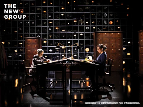 Two people sit at separate desks under lamps in a dimly lit office, surrounded by shelves of objects and filing cabinets; "THE NEW GROUP" is written in the top left corner.