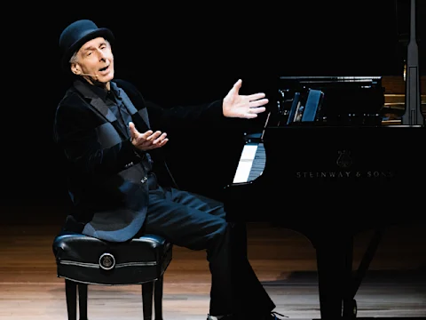 An older man in a black hat and suit sits on a piano bench with arms outstretched in front of a Steinway & Sons grand piano on stage.