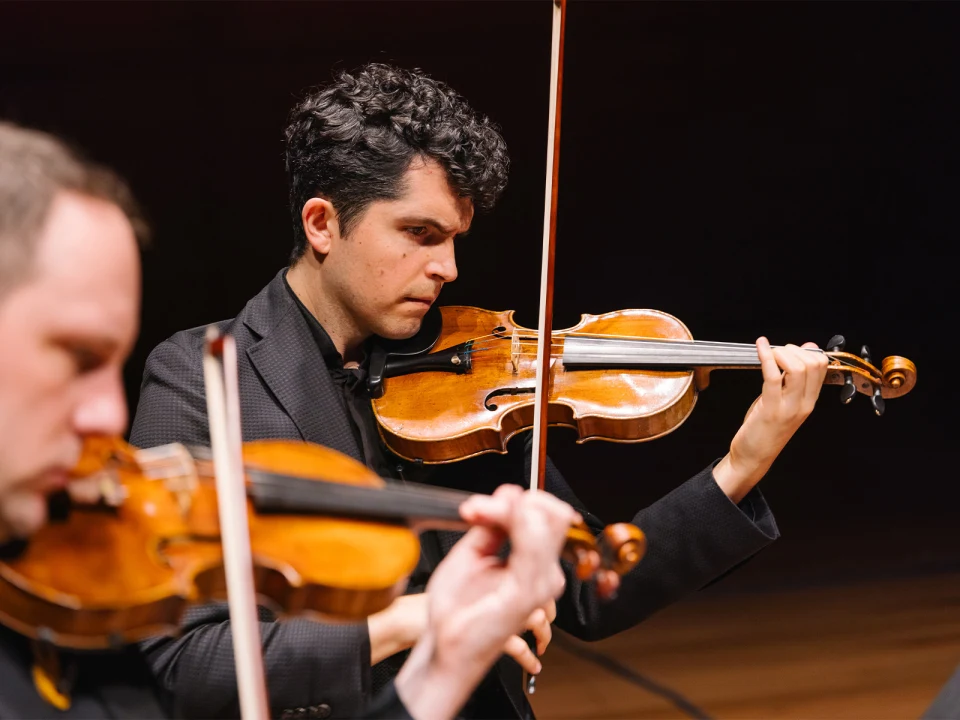 Two violinists performing on stage, focusing intently on their instruments while playing. The background is dark and the lighting highlights the musicians and their violins.