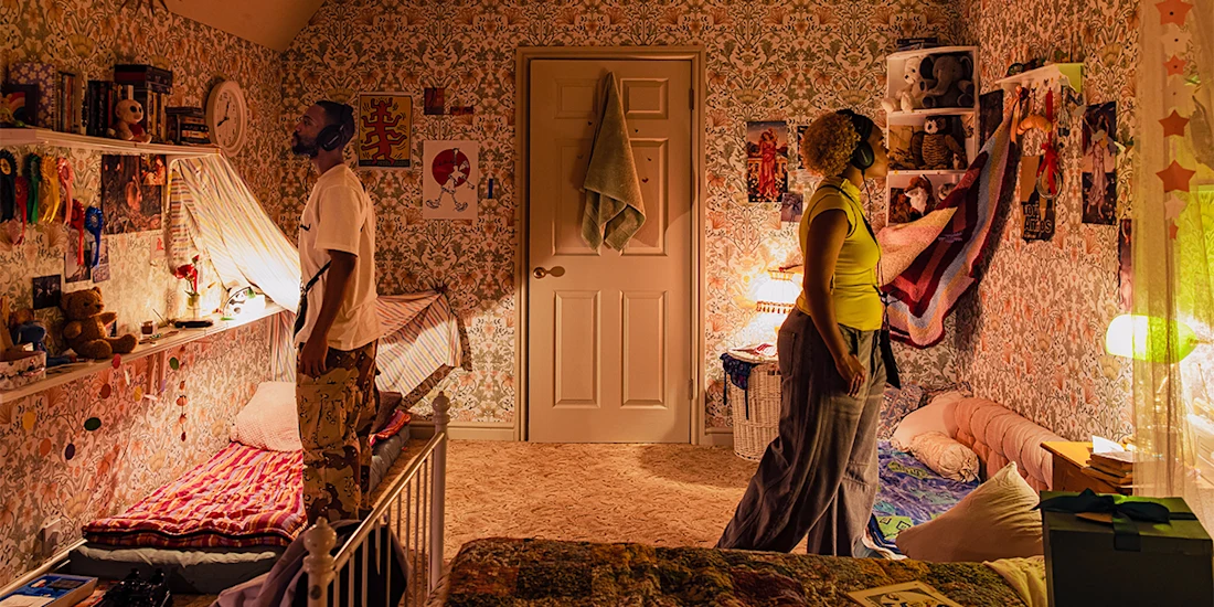 A man and a woman stand facing each other in a small, cluttered bedroom with patterned wallpaper, twin beds, and various decorations on the walls.