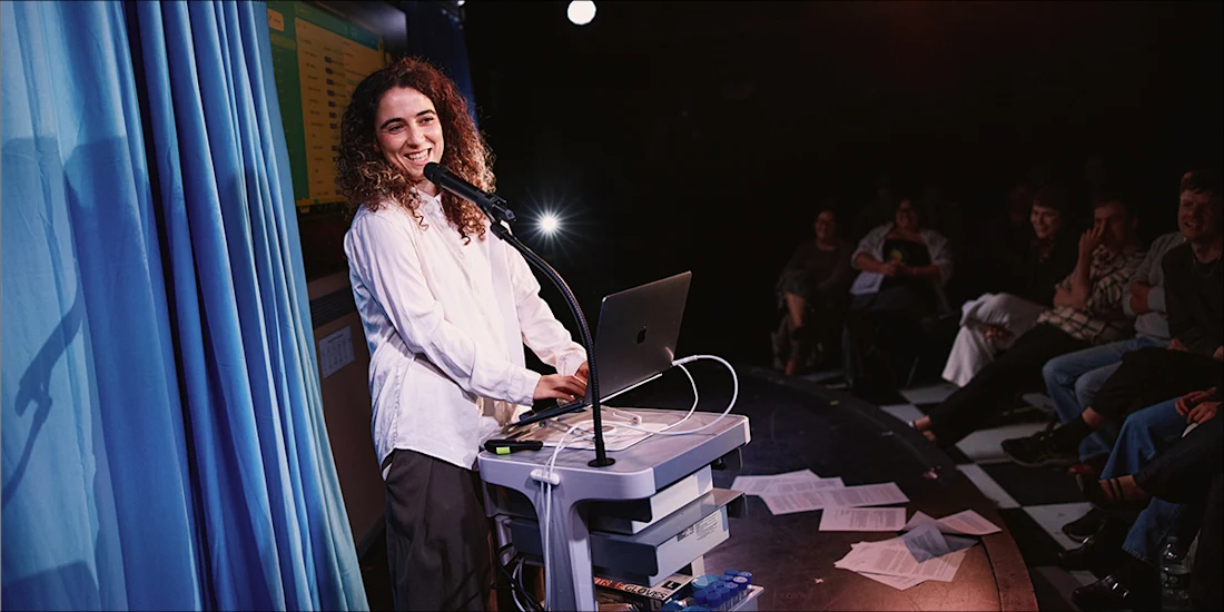 A woman with curly hair stands at a podium with a laptop, speaking into a microphone in front of an audience.