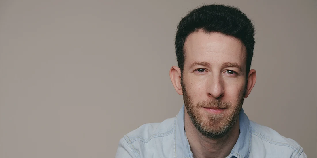 A man with short dark hair and a beard, wearing a light denim shirt, looks at the camera against a plain beige background.