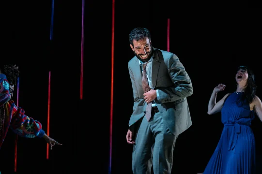 A man in a gray suit, laughing and holding his stomach, stands on a stage with two other people; one points and the other laughs in the background. Vertical lights hang behind them.