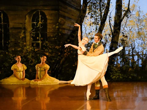 Two ballet dancers perform a duet on stage in front of a backdrop with trees and a rustic house, while two other dancers in yellow dresses sit in the background.