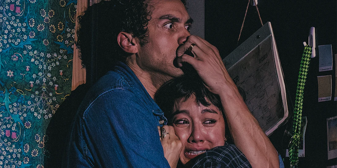 A man covers a woman’s mouth as they crouch together, both appearing frightened, in a dim room with patterned wallpaper and a whiteboard in the background.