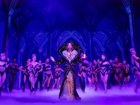 A performer in an elaborate robe stands center stage with outstretched arms, surrounded by dancers in revealing costumes, under purple lighting in a gothic-themed setting.