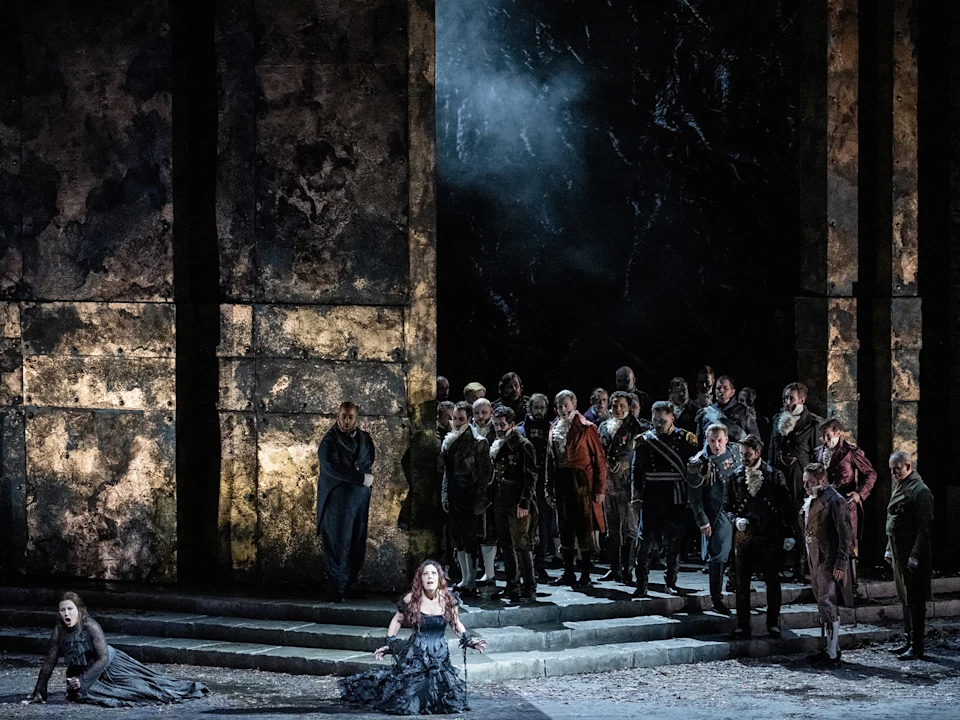 A dramatic opera scene shows a woman kneeling and singing in front of a group of people standing between tall, dark stone pillars on a dimly lit stage.
