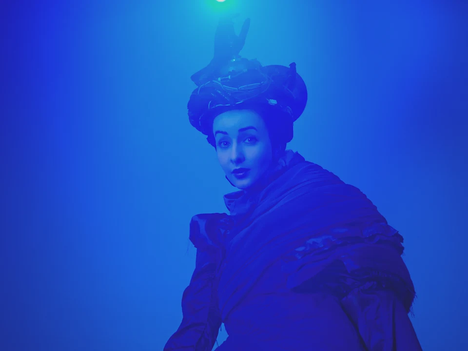 A person in dramatic lighting wears a dark, elaborate costume and headpiece, with blue light casting an ethereal glow over the scene.