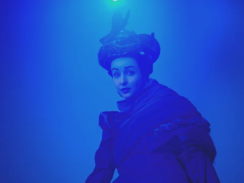 A person in dramatic lighting wears a dark, elaborate costume and headpiece, with blue light casting an ethereal glow over the scene.