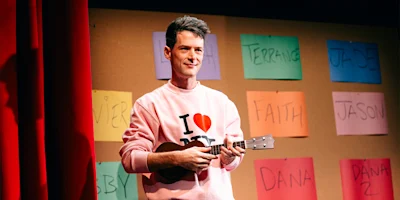 A performer in a pink “I Love NY” sweatshirt plays a ukulele on stage in front of a wall covered in colorful paper name tags.
