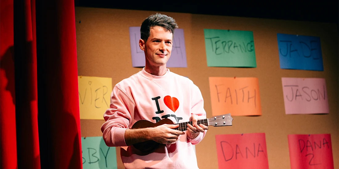 A performer in a pink “I Love NY” sweatshirt plays a ukulele on stage in front of a wall covered in colorful paper name tags.