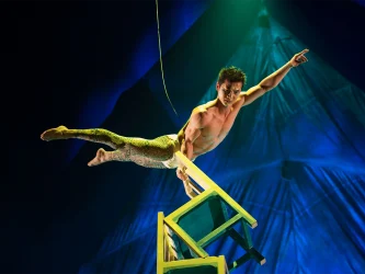 A shirtless performer balances on chairs in an acrobatic pose, with one arm pointing forward and legs outstretched. The background is dimly lit with blue and green tones.