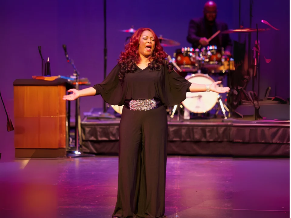 A woman in a black dress sings passionately on stage with a microphone. A drummer plays in the background, and a podium is visible to the left.