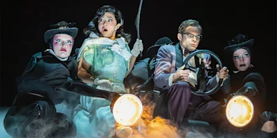 Actors on stage perform a dramatic car scene with exaggerated expressions, two holding headlights, others dressed in black with theatrical makeup, and one person gripping a steering wheel.
