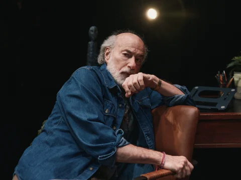 A bearded man in a denim shirt sits pensively in front of a desk, with a spotlight shining behind him.