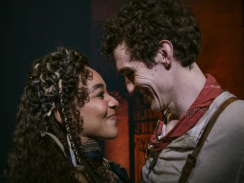 Two people facing each other closely, smiling, and appearing happy. Both have curly hair; the individual on the left has braids, and the one on the right wears a red bandana.