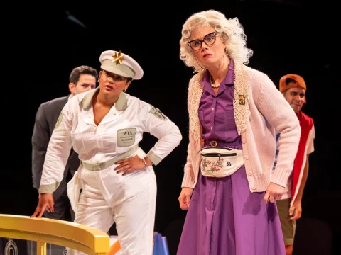 Four actors perform on stage. One wears a white uniform, another has a white wig and glasses, dressed in purple with a fanny pack. Another actor is partly visible, dressed in red and white.