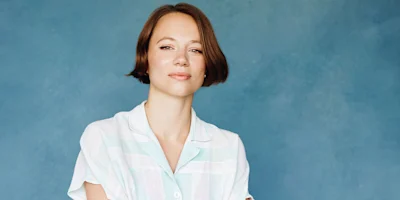 A woman with a short bob haircut wearing a light-colored, short-sleeved collared shirt against a blue background.